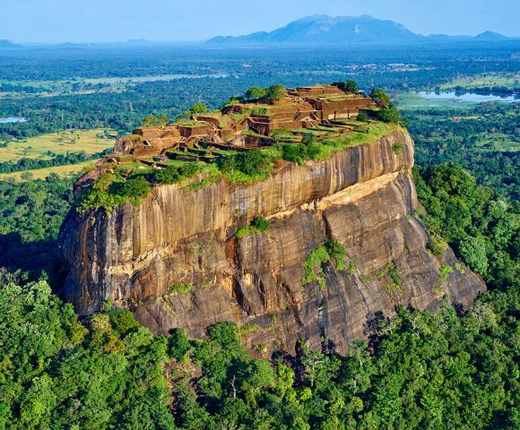 Aerial view of Sigiriya, Sri Lanka, showcasing the ancient rock fortress atop a massive rock formation, surrounded by lush green forest and distant mountains.