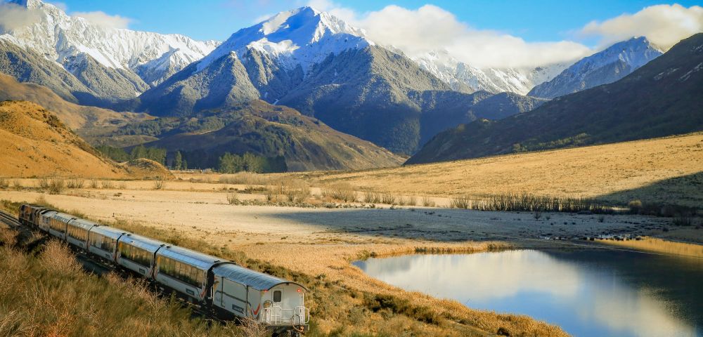 A passenger train travels through a wide valley with golden grasslands beside a reflective lake, set against a backdrop of tall, rugged mountains covered in snow under a partly cloudy sky.