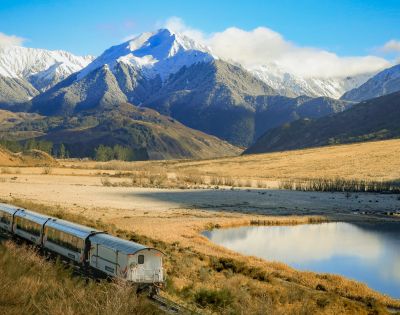 A passenger train travels through a wide valley with golden grasslands beside a reflective lake, set against a backdrop of tall, rugged mountains covered in snow under a partly cloudy sky.
