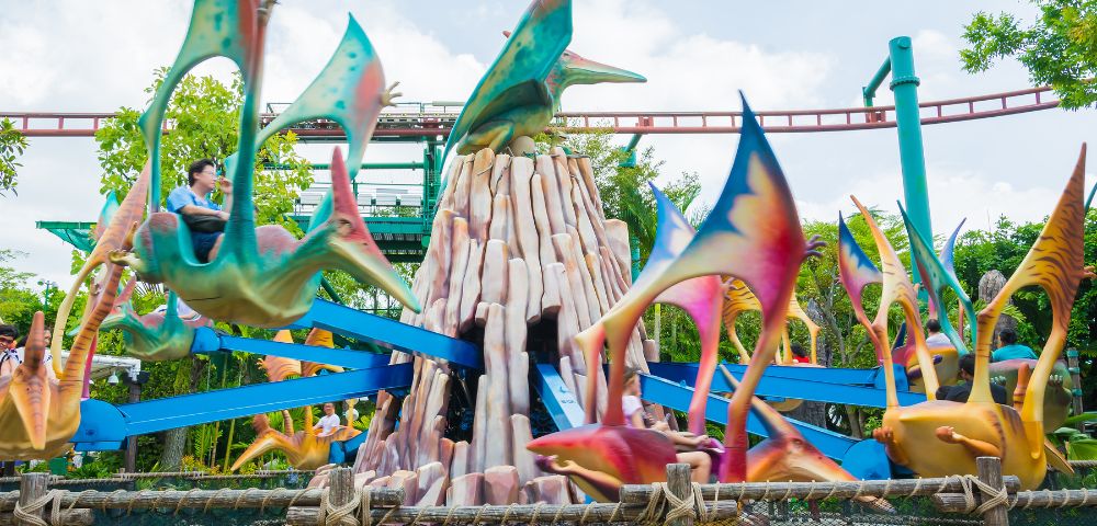 A colorful amusement park ride featuring flying dinosaur‑shaped vehicles rotates around a rocky central structure, with track rails and lush greenery visible in the background under a bright sky.
