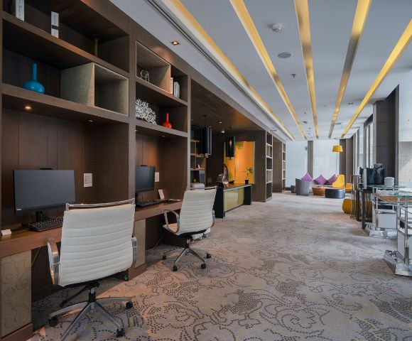 Modern office space with two white chairs and computers on wooden desks. Warm lighting, patterned carpet, and colorful bean bags create a relaxed atmosphere.