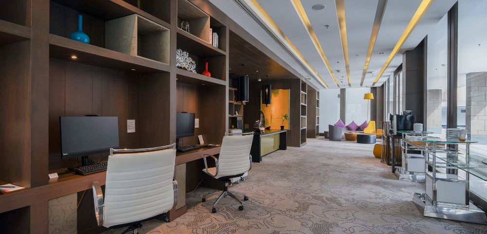 Modern office space with two white chairs and computers on wooden desks. Warm lighting, patterned carpet, and colorful bean bags create a relaxed atmosphere.