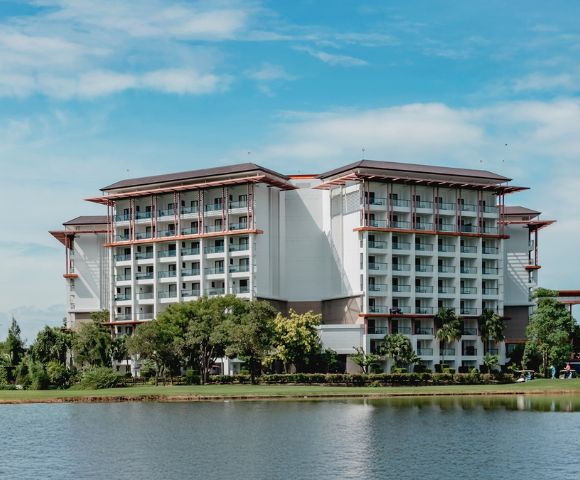 A modern nine-story hotel with balconies, surrounded by trees, sits beside a calm lake under a bright blue sky with scattered clouds.