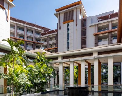 Modern resort courtyard with tropical plants, a water feature, and multi-story buildings with wooden accents under a clear blue sky. Calm and inviting atmosphere.