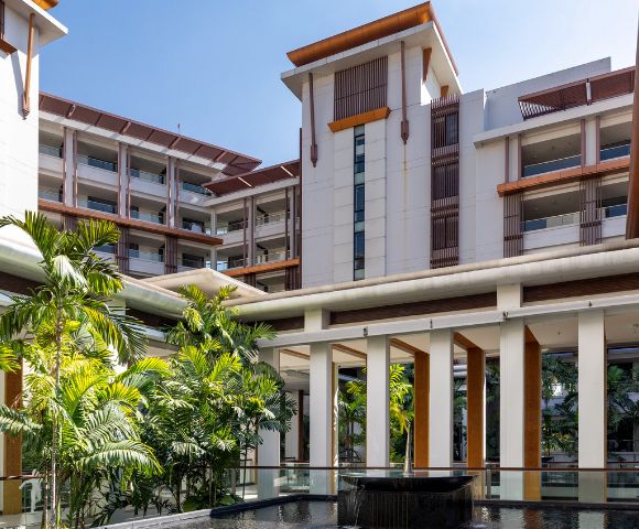 Modern resort courtyard with tropical plants, a water feature, and multi-story buildings with wooden accents under a clear blue sky. Calm and inviting atmosphere.