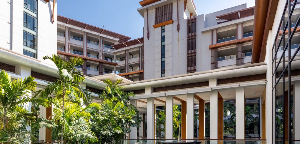 Modern resort courtyard with tropical plants, a water feature, and multi-story buildings with wooden accents under a clear blue sky. Calm and inviting atmosphere.