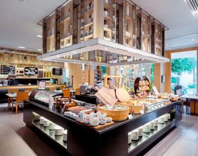 Modern buffet restaurant with diverse food spread. Elegant wooden shelves and bright lighting create a welcoming atmosphere. Tables visible in the background.