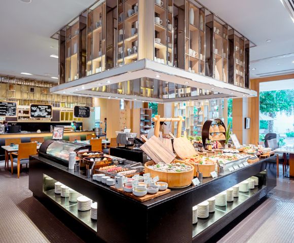 Modern buffet restaurant with diverse food spread. Elegant wooden shelves and bright lighting create a welcoming atmosphere. Tables visible in the background.
