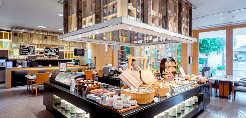 Modern buffet restaurant with diverse food spread. Elegant wooden shelves and bright lighting create a welcoming atmosphere. Tables visible in the background.