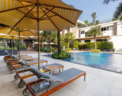Outdoor pool area with sun loungers and large striped umbrellas on a sunny day. Modern building, lush greenery, and a tranquil atmosphere.