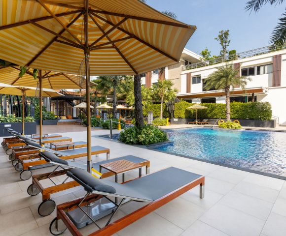 Outdoor pool area with sun loungers and large striped umbrellas on a sunny day. Modern building, lush greenery, and a tranquil atmosphere.