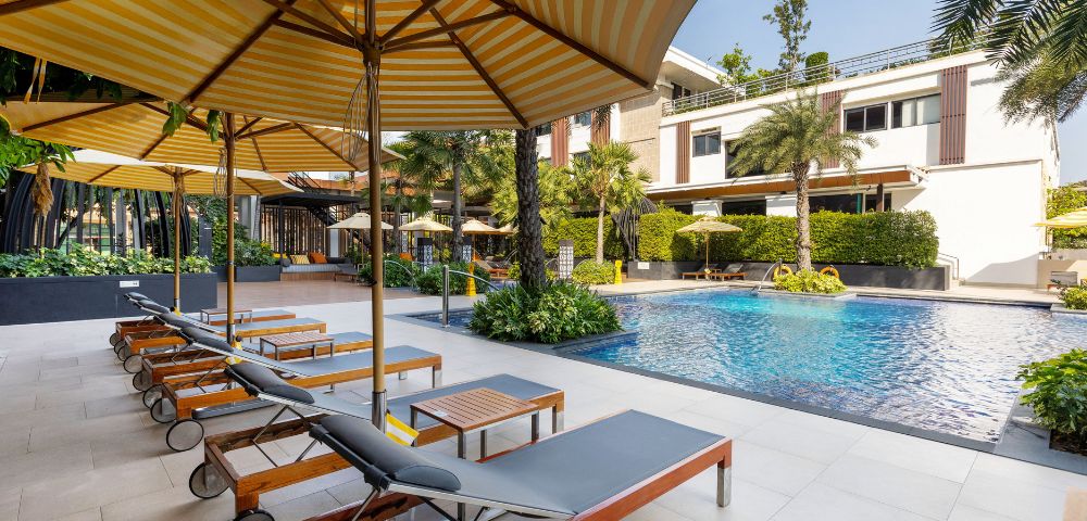 Outdoor pool area with sun loungers and large striped umbrellas on a sunny day. Modern building, lush greenery, and a tranquil atmosphere.