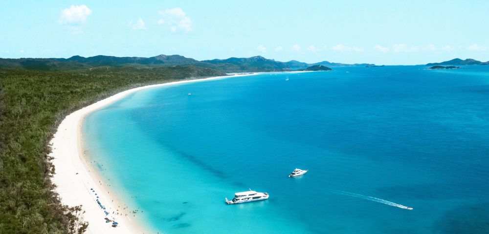 Aerial view of a serene beach with lush green hills, turquoise water, and two boats. The scene conveys tranquility and natural beauty.