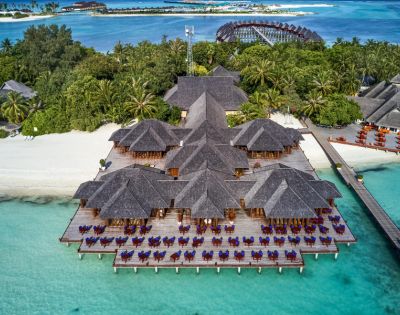 Aerial view of a tropical resort with beachfront bungalows, surrounded by lush greenery and turquoise waters under a clear blue sky.