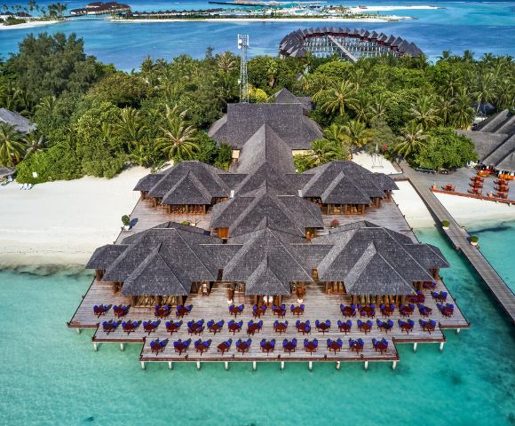 Aerial view of a tropical resort with beachfront bungalows, surrounded by lush greenery and turquoise waters under a clear blue sky.