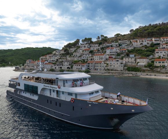A luxurious yacht sails along a coastal town with tiled roofs and lush greenery under a partly cloudy sky, conveying a serene and picturesque scene.