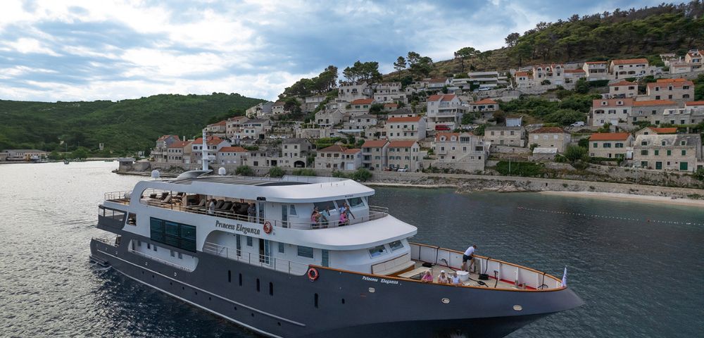 A luxurious yacht sails along a coastal town with tiled roofs and lush greenery under a partly cloudy sky, conveying a serene and picturesque scene.