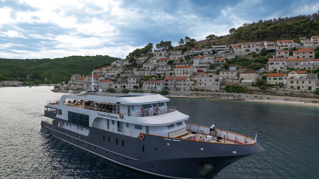 A luxurious yacht sails along a coastal town with tiled roofs and lush greenery under a partly cloudy sky, conveying a serene and picturesque scene.