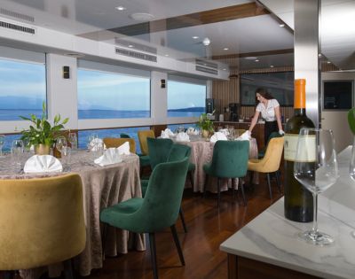 Elegant dining room on a boat with round tables set for a meal, colorful chairs, and a staff member arranging a table. Large windows offer a sea view.