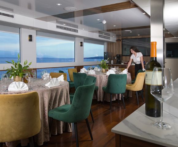 Elegant dining room on a boat with round tables set for a meal, colorful chairs, and a staff member arranging a table. Large windows offer a sea view.