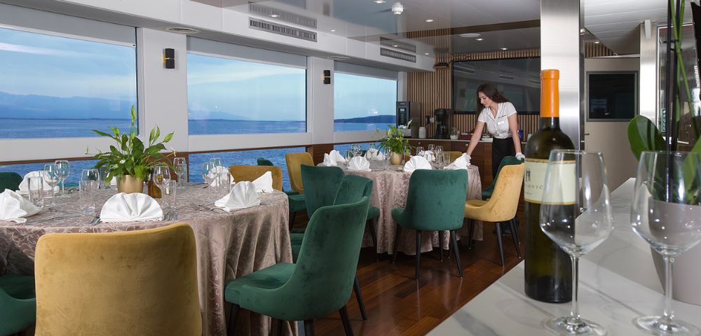 Elegant dining room on a boat with round tables set for a meal, colorful chairs, and a staff member arranging a table. Large windows offer a sea view.