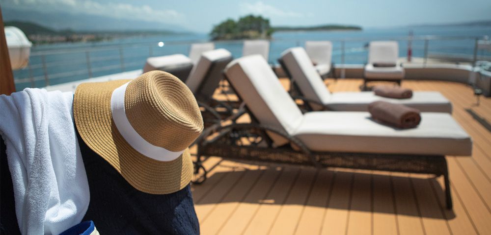 Sunlit deck with white loungers facing the sea, surrounded by blue skies. A straw hat and towel hang over a chair, creating a relaxed, vacation vibe.