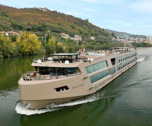 A sleek, modern river cruise ship sails along a calm river, surrounded by lush green hills and quaint hillside houses under a clear blue sky.