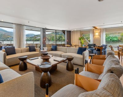 Spacious, modern living room with beige sofas, wooden tables, and blue and brown accents. Large windows reveal scenic mountain and river views.