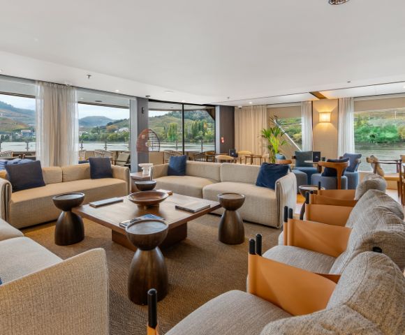 Spacious, modern living room with beige sofas, wooden tables, and blue and brown accents. Large windows reveal scenic mountain and river views.