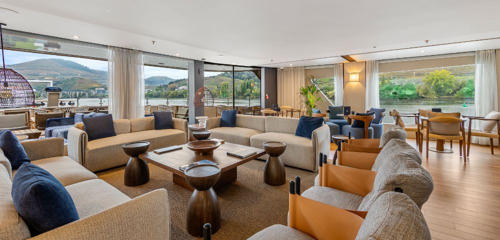 Spacious, modern living room with beige sofas, wooden tables, and blue and brown accents. Large windows reveal scenic mountain and river views.