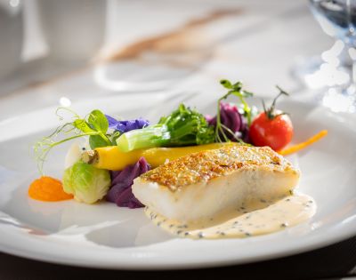 A gourmet dish features a seared fish fillet on creamy sauce, garnished with colorful vegetables and a cherry tomato, arranged artfully on a white plate.