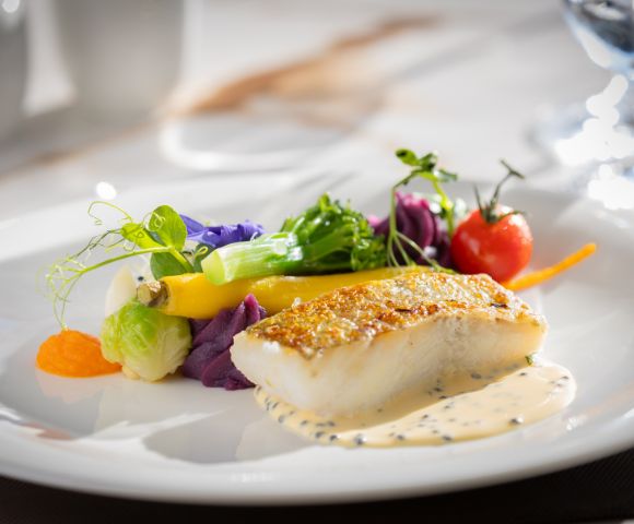 A gourmet dish features a seared fish fillet on creamy sauce, garnished with colorful vegetables and a cherry tomato, arranged artfully on a white plate.
