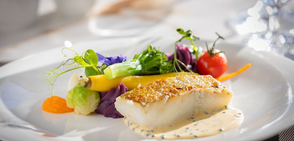 A gourmet dish features a seared fish fillet on creamy sauce, garnished with colorful vegetables and a cherry tomato, arranged artfully on a white plate.