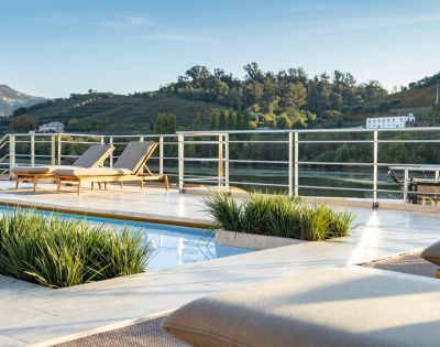 Luxurious poolside scene with two loungers on a sunlit deck overlooking lush, green hills and a serene river. Relaxing, tranquil atmosphere.