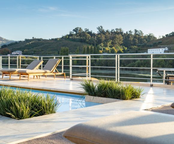 Luxurious poolside scene with two loungers on a sunlit deck overlooking lush, green hills and a serene river. Relaxing, tranquil atmosphere.