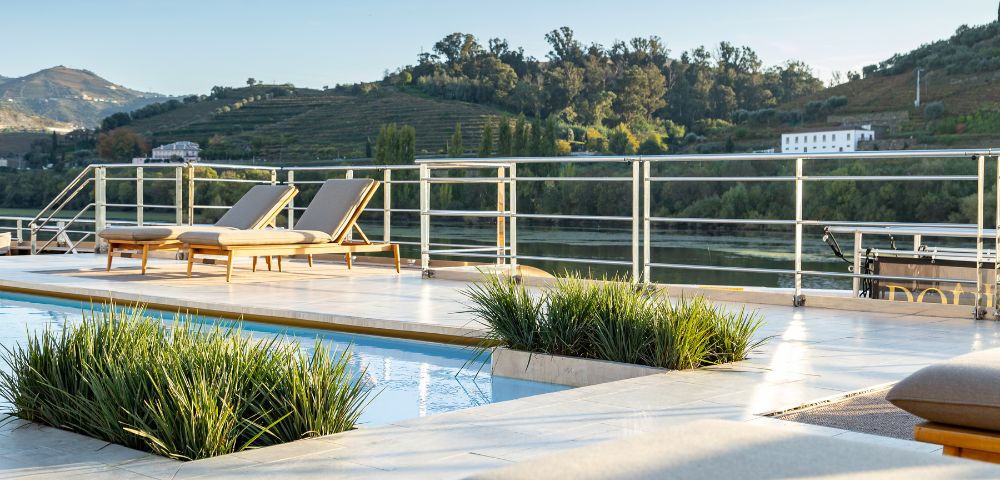 Luxurious poolside scene with two loungers on a sunlit deck overlooking lush, green hills and a serene river. Relaxing, tranquil atmosphere.