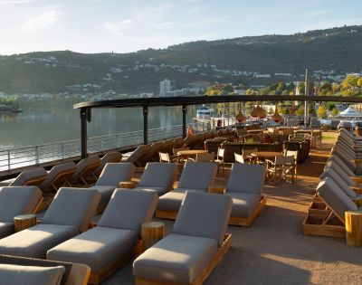 Riverside outdoor lounge with rows of wooden loungers and chairs under a shaded pergola, overlooking a calm river and distant green hills at sunset. Calm atmosphere.