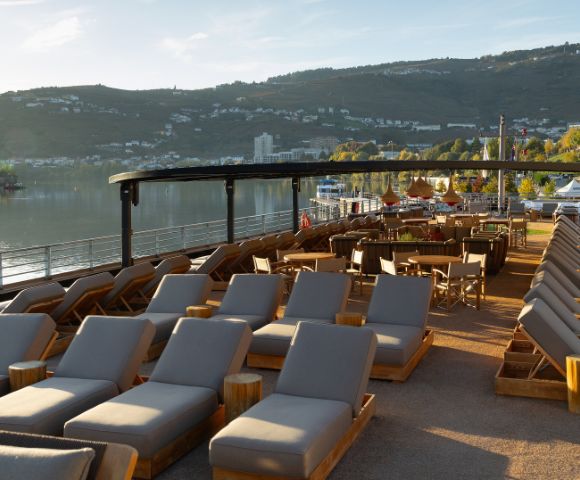 Riverside outdoor lounge with rows of wooden loungers and chairs under a shaded pergola, overlooking a calm river and distant green hills at sunset. Calm atmosphere.