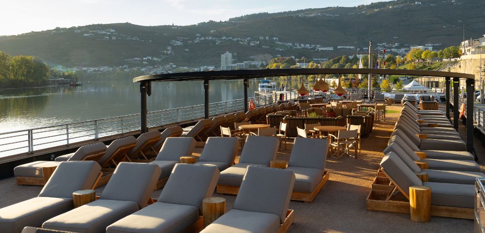 Riverside outdoor lounge with rows of wooden loungers and chairs under a shaded pergola, overlooking a calm river and distant green hills at sunset. Calm atmosphere.