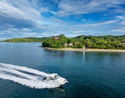 A speedboat cuts through tranquil blue waters near a lush, tropical island with green hills and scattered beach huts, under a partly cloudy sky.
