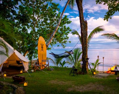 Tropical beach glamping scene with a tent, surfboard, hammock, and a fire pit at sunset. Palm trees and lanterns create a serene, cozy atmosphere.