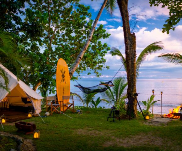 Tropical beach glamping scene with a tent, surfboard, hammock, and a fire pit at sunset. Palm trees and lanterns create a serene, cozy atmosphere.