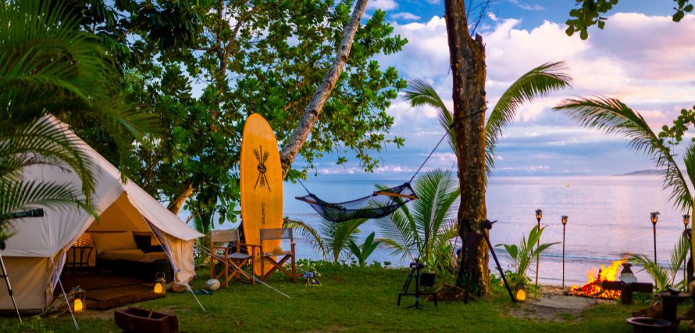 Tropical beach glamping scene with a tent, surfboard, hammock, and a fire pit at sunset. Palm trees and lanterns create a serene, cozy atmosphere.