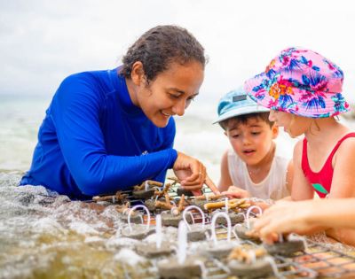 A person in a blue shirt helps two children with coral planting in shallow water. The kids are focused, one in a floral hat, conveying a sense of learning and conservation.