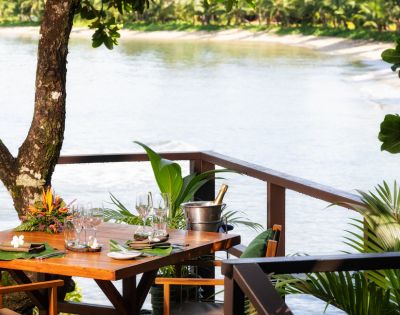 A wooden table set with glasses and a wine bucket overlooks a serene river. Lush greenery surrounds the deck, conveying a tranquil, tropical atmosphere.