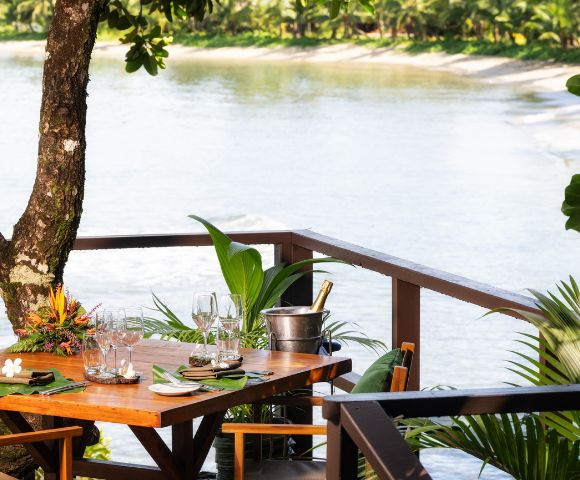 A wooden table set with glasses and a wine bucket overlooks a serene river. Lush greenery surrounds the deck, conveying a tranquil, tropical atmosphere.