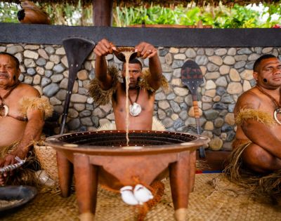 Three men in traditional attire prepare kava in a large wooden bowl. The man in the center pours liquid, with stone walls and foliage in the background.