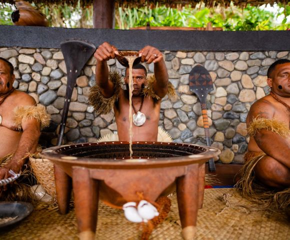 Three men in traditional attire prepare kava in a large wooden bowl. The man in the center pours liquid, with stone walls and foliage in the background.