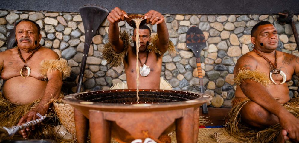 Three men in traditional attire prepare kava in a large wooden bowl. The man in the center pours liquid, with stone walls and foliage in the background.