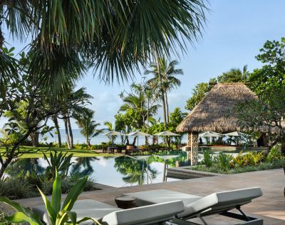 Tropical resort scene with palm trees, a tranquil pool, and a thatched gazebo. Two white sun loungers in the foreground suggest a relaxing vibe.